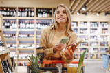 Fototapeta Na sufit - Young minded woman wear brown shirt casual clothes hold wine use mobile cell phone shopping at supermaket store with trolley cart buy choose products inside hypermarket. Purchasing gastronomy concept.