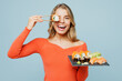 Young happy woman wear orange casual clothes hold cover eye with bar of eat raw fresh sushi roll served on black plate Japanese food with chopsticks isolated on plain blue background studio portrait.