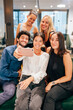 © carlesmiro - Young creative startup team smiling and taking selfie on mobile phone while gathering together in modern workspace. Blurred group of people.
