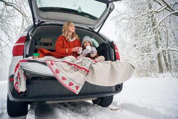 Drinking from the cup. Woman with her daughter is on the trunk of a car in the winter meadow and forest