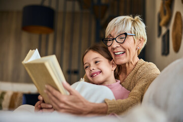 Wall Mural - A smiling grandchild spending time with her grandma, reading a book together, covered in a blanket.