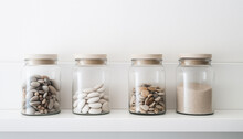 Jars Containing Beach Pebbles And Sand On White Cupboard. Concept Of Preserving Good Summer Memories. Minimalistic Wallpaper. 