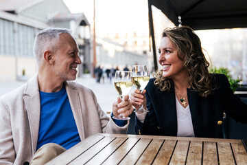 Lovely mid adult couple toasting with wine while sitting in a terrace restaurant. Romantic date of a middle age woman and man drinking with glasses at a bar terrace looking each other.