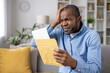 Senior gray haired man upset received envelope mail message notification with bad news, sad displeased and disagreeable african american man sitting at home on sofa inside living room.