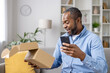 © Liubomir - Happy and satisfied buyer received cardboard box in mail, satisfied with speed of delivery of a gift for an online store, an African American man with a phone in hands confirms receipt of the goods.
