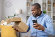 Dissatisfied sad online buyer, man received wrong damaged parcel, african american man sitting at home on sofa in living room with cardboard box in hands and phone, writing negative review.