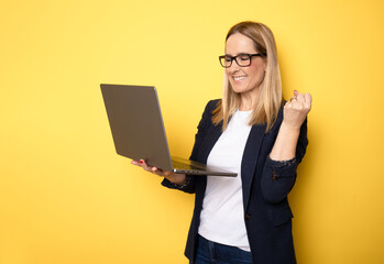 Smiling cheerful funny beautiful attractive young woman 40s wearing basic casual jacket standing working on laptop pc computer isolated on yellow colour background studio portrait