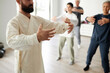 © Seventyfour - Cropped shot of bearded male qigong master teaching qigong tree pose, students repeating standing meditation exercise in blurred background