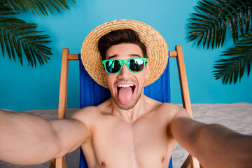 Wall Mural - Photo of cheerful carefree guy swimwear sunhat relaxing lounge chair stick out vlogging recording video isolated blue color background