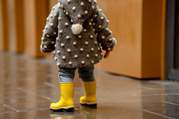 Wall Mural - A cute stylish two-year-old toddler kid girl in a gray coat and yellow boots walks outdoors near buildings with glass walls