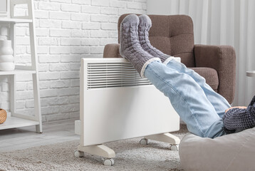 Poster - Young woman in warm winter socks near electric heater. Concept of heating season