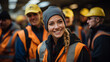 A joyful woman stands among a group of people wearing bright orange and yellow vests, her smile radiating as she confidently dons a hard hat on her head