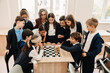 © Drobot Dean - Group of kids and teacher watching chess game between two boys while standing around table in classroom