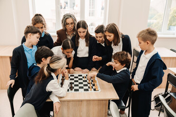 Group of kids and teacher watching chess game between two boys while standing around table in classroom