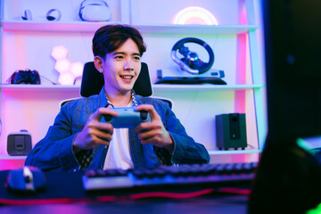 Young positive happy Asian man holding and pressing buttons of a wireless controller playing a video game in a bedroom with red and blue neon lights at home. Leisure time and esports gamer concept