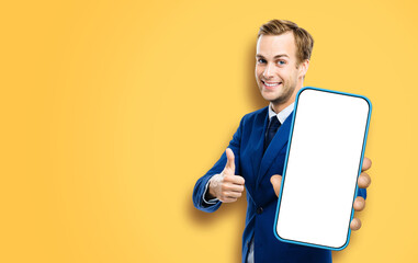 Excited smiling young business man showing cell phone, mobile smartphone. Happy businessman hold cellphone white screen, isolated against orange yellow color background. Thumb up. Great app ad concept