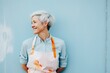 A confident and attractive elderly woman artist against a blue wall in an apron, personifying positivity, leading a stylish and active lifestyle in her 60s.
