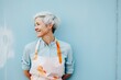 © Iryna - A confident and attractive elderly woman artist against a blue wall in an apron, personifying positivity, leading a stylish and active lifestyle in her 60s.