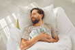© Studio Romantic - Rich man sleeps with cash in hands. Bearded man lying on white pillow, holding dollar bills on his chest and dreaming sweet dreams with happy, peaceful face expression. Money, finance, savings concept