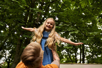 tattooed man holding and playing with cheerful daughter in green park, happy family moments