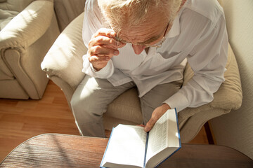 Wall Mural - Lifestyle portrait of senior man reading Holy Bible at home. Natural aesthetic light