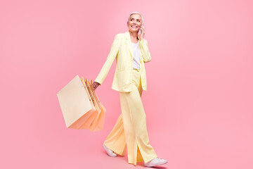 Poster - Full size photo of adorable senior woman go to empty space holding shopping bags talk on smartphone isolated on pink color background