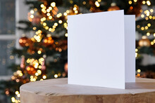 Standing Blank Empty Square Greeting Card Mock Up Before A Blurred Christmas Tree With Copy Space. For Use As A Christmas, Background Template.    