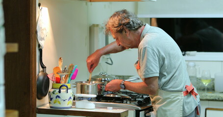 Wall Mural - Senior man cooking at the kitchen. Candid older retired person stirring pot