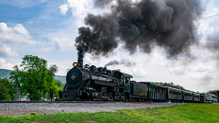Canvas Print - A View of a Narrow Gauge Restored Steam Passenger Train Blowing Smoke, Starting To Pull Out of a Station on a Summer Day