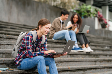 Happy diverse university students sitting on steps, using laptops and tablets, enjoying conversation. Student exchange and study abroad program, Asian man, african american woman and caucasian people.