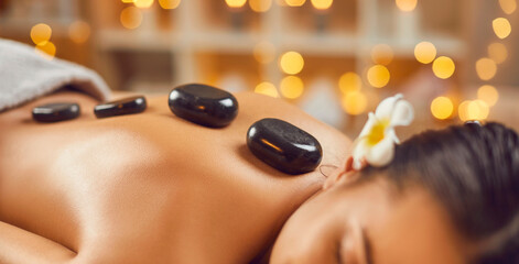 Wall Mural - Close up photo of female back with hot stones. Portrait of pretty young brunette woman with closed eyes lying alone relaxing in spa salon getting massage therapy. Wellness and beauty day concept.