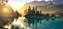 Idyllic Lake At Sunrise, A Picturesque Panorama With Majestic Mountains And The Golden Sunlight Coming In Behind The Trees