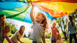 © Gautier Normand - Un groupe d'enfants souriants, jouant à l'extérieur avec un grand parachute multicolore, en colonie de vacances lors d'une journée ensoleillée.