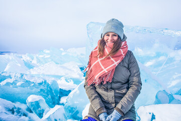 Smiling joyful female wanderer looking in camera while sitting on frozen Baikal lake against large cracked ice floes during winter vacation 