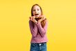 Portrait of attractive excited little girl wearing casual clothes, eating tasty pink donut, standing isolated on yellow background. Concept of food, dessert