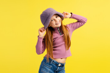 Wall Mural - Portrait of smiling beautiful little girl wearing stylish purple hat and casual clothes looking at camera, model posing isolated on yellow background. Fashion concept