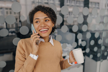Wall Mural - Smiling female manager with coffee cup is talking phone while standing on modern office background