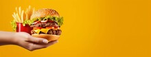 Burger And Fries Menu On Christmas Day. Hand Holding A Delicious Big Cheeseburger With Fries And Ketchup Sauce On A Yellow Color Background, Festive Christmas And New Year Theme, For Posters And Banne