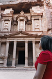 Fototapeta Na drzwi - Young European tourist looking at the Treasury of Petra, Jordan. Brunette girl in red dress enjoying the view of the façade and the ruins of the ancient city carved in stone.