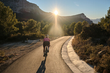 Fit female cyclist wearing cycling kit and helmet riding on the road on a gravel bike at sunset.Empty mountain road. Sports motivation image.Calpe town in Spain.