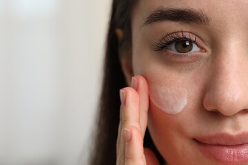 Poster - Young woman with dry skin applying cream onto her face on blurred background, closeup. Space for text