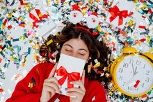 Girl Hiding Face With Gift Box And Lying On Confetti Near Clock