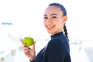 Wall Mural - Young moroccan girl with an apple at outdoors smiling a lot