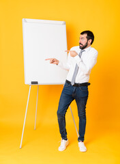 Wall Mural - Full-length shot of businessman giving a presentation on white board over isolated yellow background frightened and pointing to the side