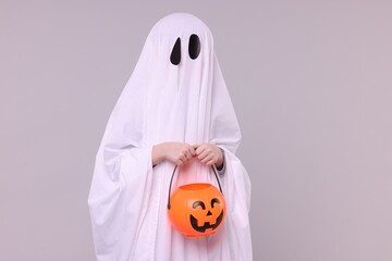 Wall Mural - Child in white ghost costume holding pumpkin bucket on light grey background. Halloween celebration