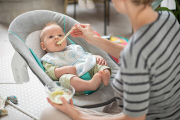 Wall Mural - Mother spoon feeding her baby boy child in baby chair with fruit puree. Baby solid food introduction concept