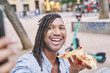 attractive man with braids holding a slice of pizza in his hand while taking a photo with his smart phone on the street