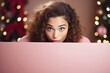 Young woman peeking out from behind a white board with a defocused Christmas background. Cozy atmosphere, pink Christmas concept. Christmas promotional banner mockup with copy space.