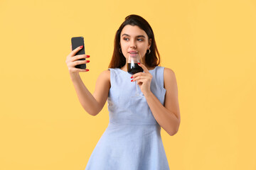 Wall Mural - Young woman with glass of wine and mobile phone video chatting on yellow background. Online dating concept