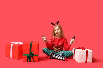 Sticker - Cute little girl in reindeer horns with chocolate Santa Claus and gift boxes on red background